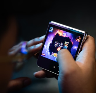 A person is holding a small electronic device, possibly a smartphone with a foldable screen, playing a video game. The game features a dark, fiery theme with icons or characters displayed on the screen.